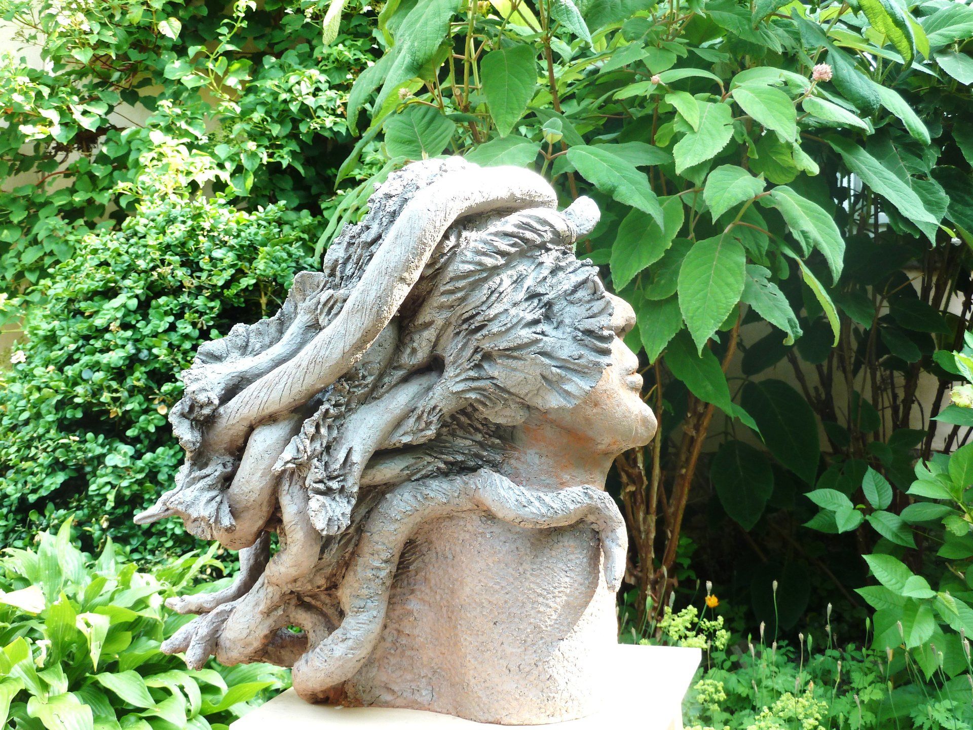 Steinbüste einer Figur mit wallendem Haar in einer üppig grünen Gartenlandschaft
