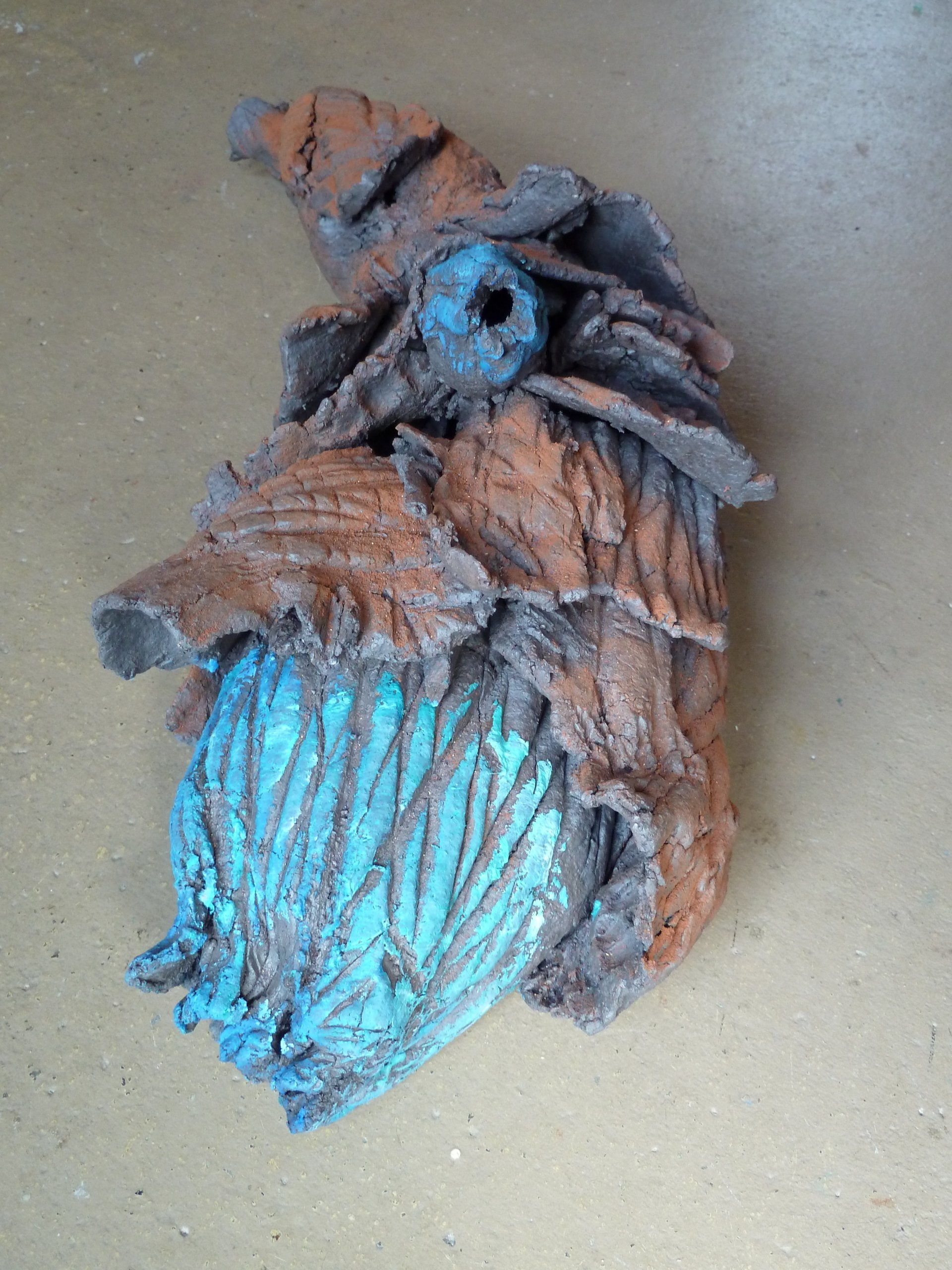 Nahaufnahme einer braun-türkisfarbenen, geschnitzten Figur auf einem schlichten, hellbraunen Untergrund.