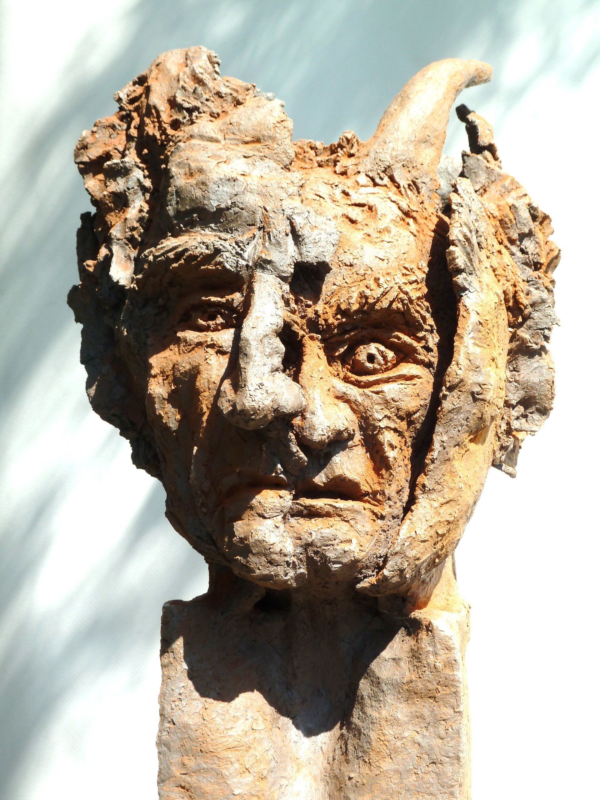 Verwitterte Bronzebüste einer gehörnten Figur mit strengem Gesicht, vom Sonnenlicht vor hellem Himmel beleuchtet