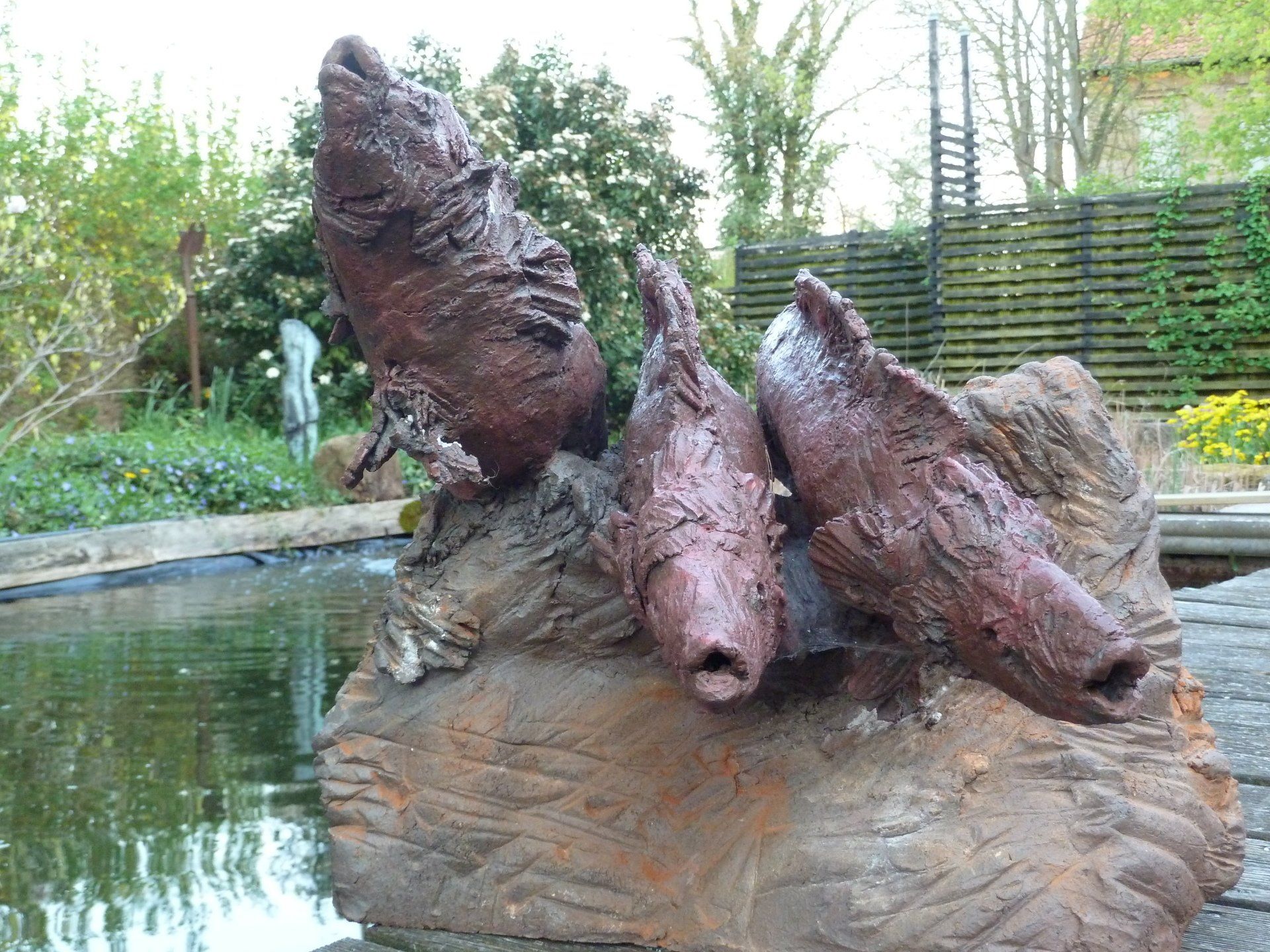 Rostige abstrakte Skulptur neben einem Teich, eingebettet in einen grünen Garten mit Bambuszaun.