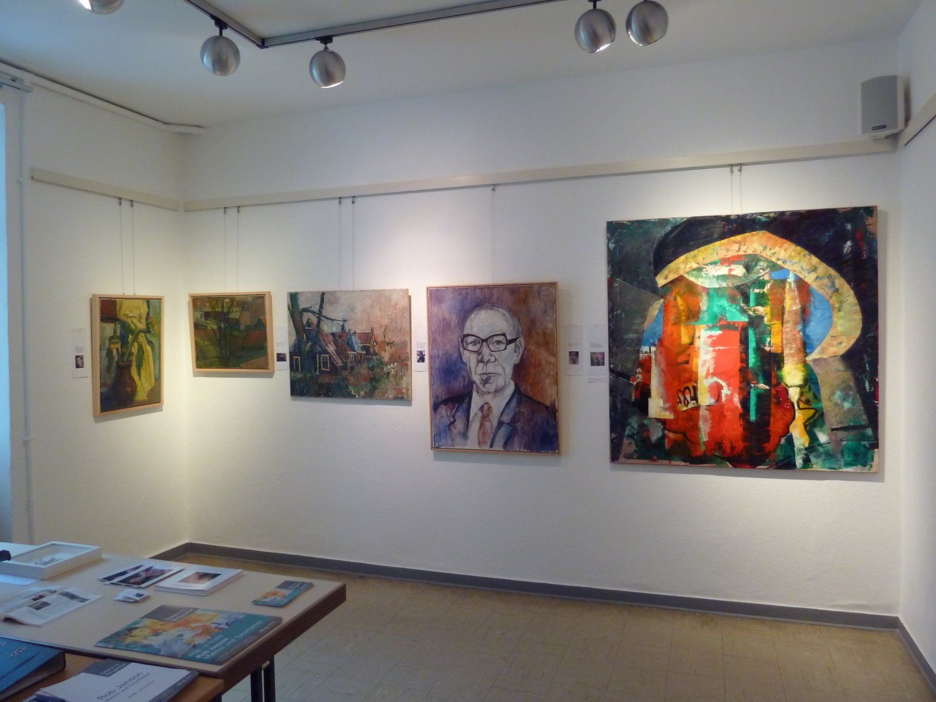 Kunstgaleriewand mit fünf farbenfrohen Gemälden unter Schienenbeleuchtung, vom anderen Ende des Raumes aus betrachtet.
