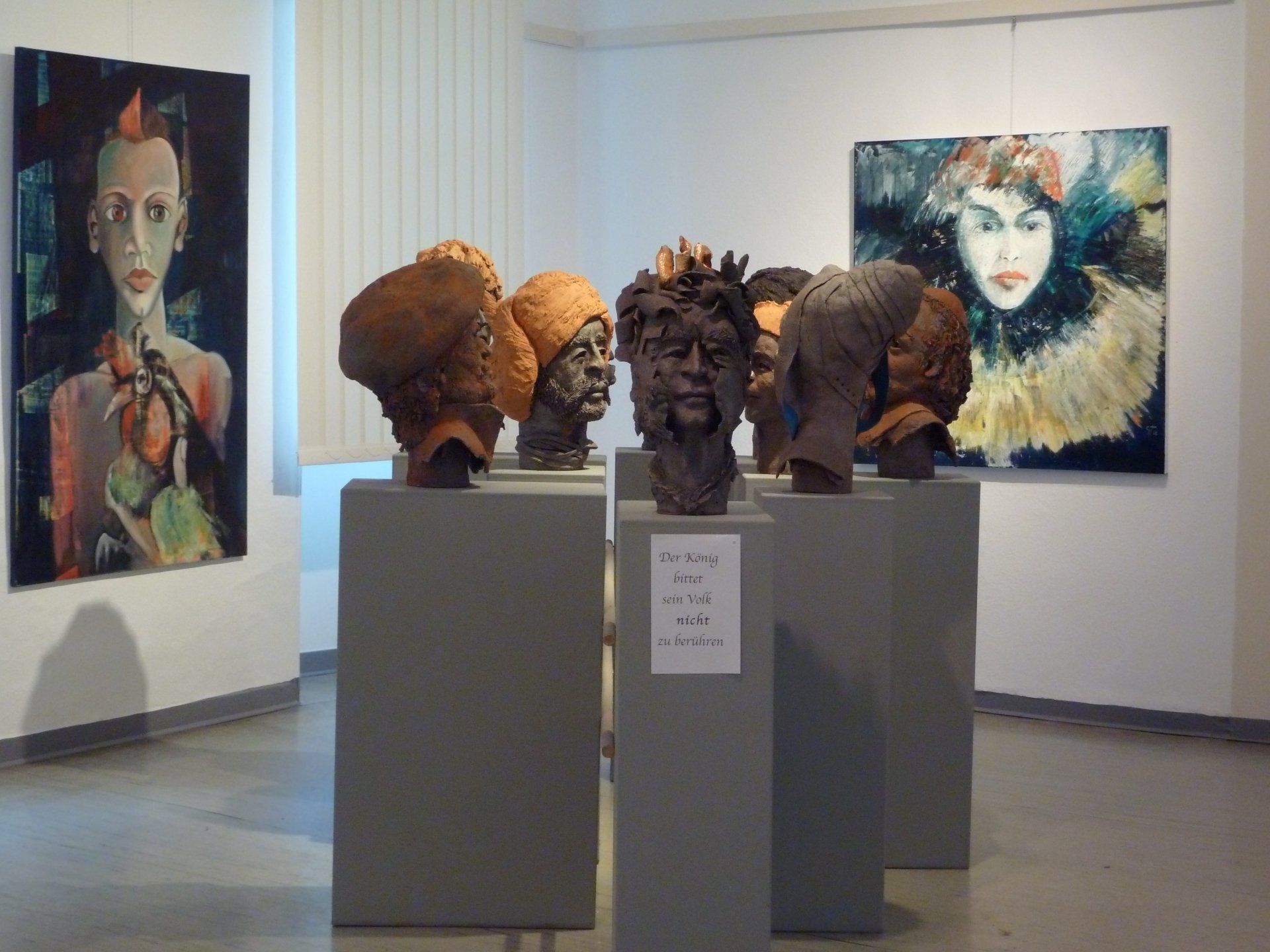 Ausstellungsstück in einer Kunstgalerie: vier Skulpturenköpfe auf Sockeln und zwei farbenfrohe Porträtgemälde an den Wänden.