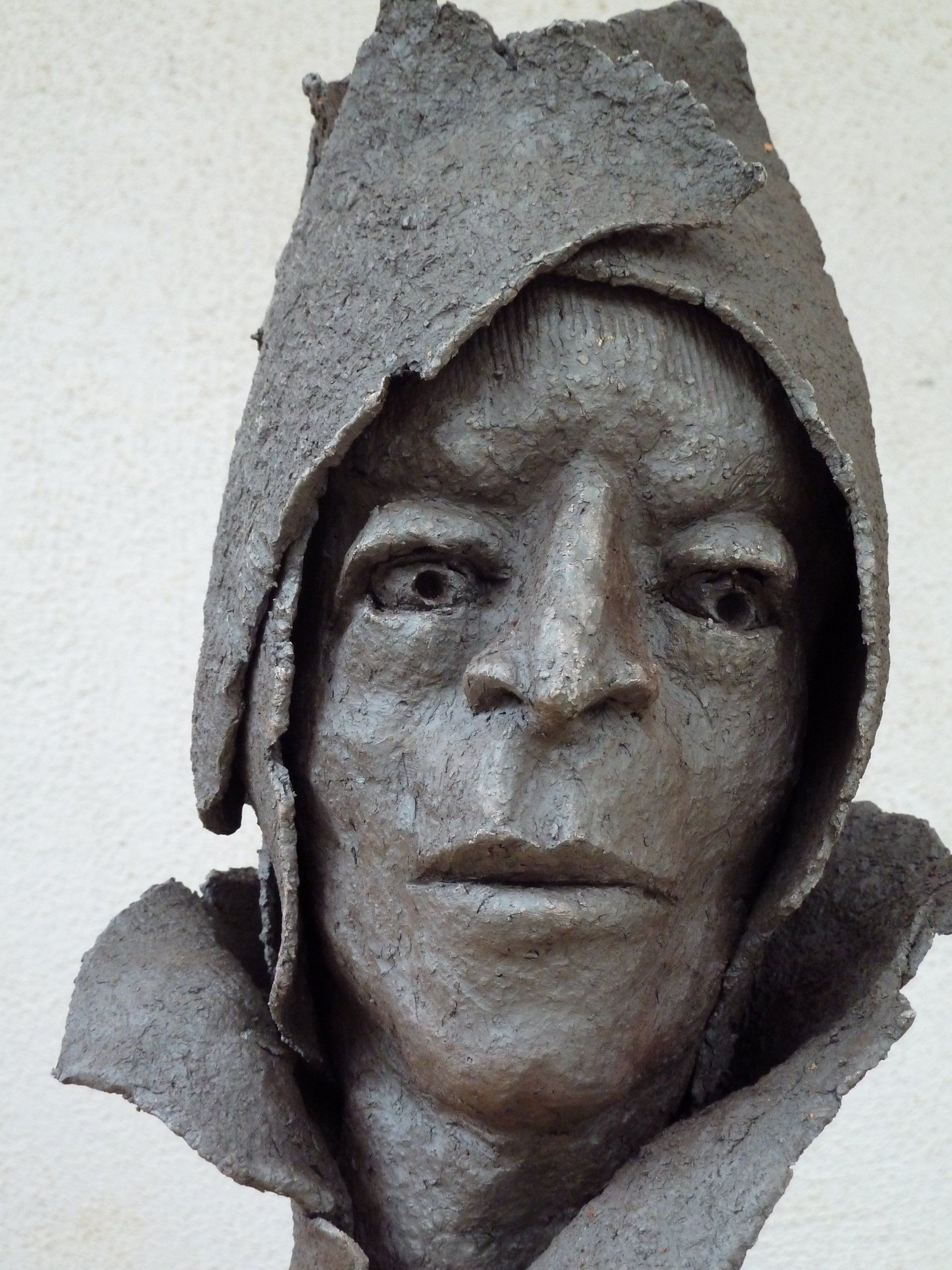 Verwitterte Stein- oder Tonskulptur eines Gesichts mit Kapuze, das mit neutralem Gesichtsausdruck nach vorn blickt