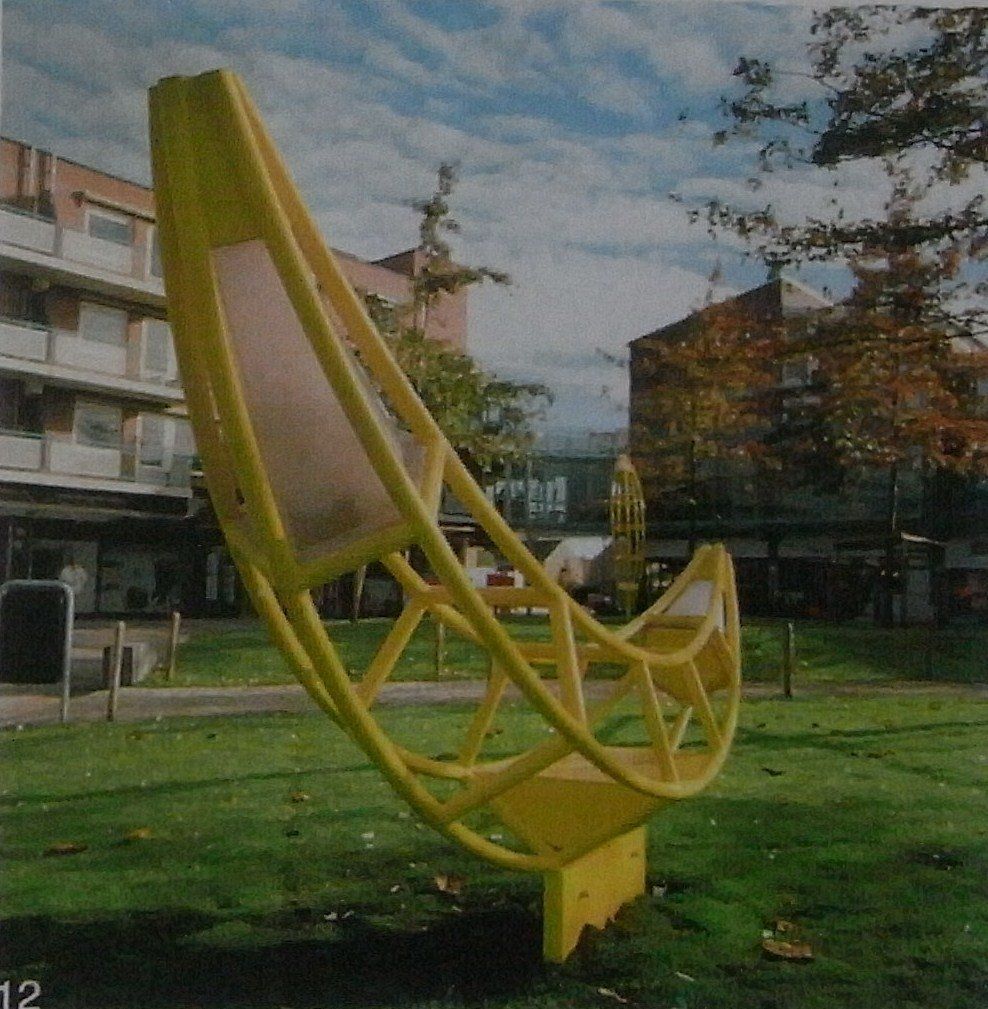 Gelbe, gebogene Stuhlskulptur auf einer Rasenfläche vor Wohnhäusern
