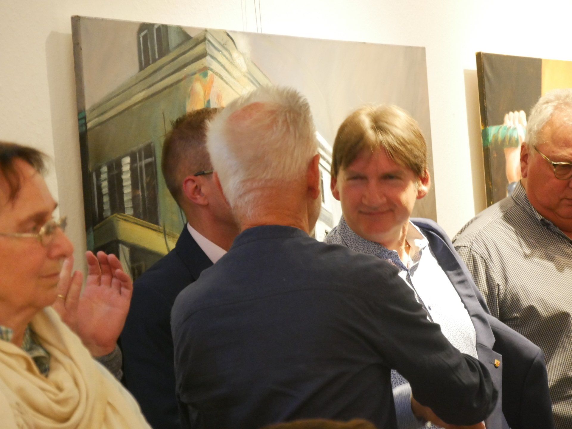 Eine kleine Gruppe unterhält sich drinnen neben einem Wandbild; eine Person gestikuliert, während die anderen zuhören und lächeln.
