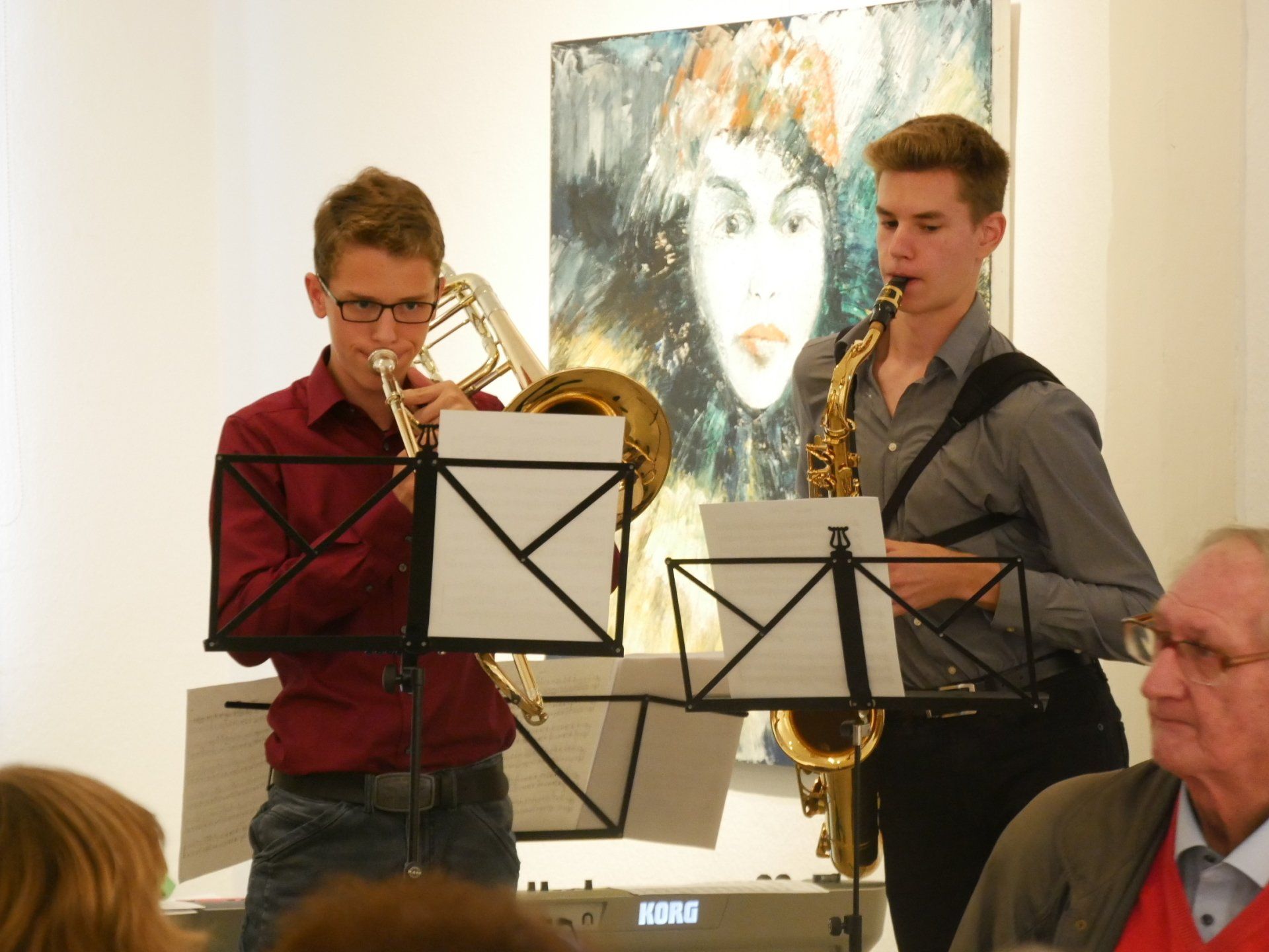 Zwei Musiker spielen in einem Innenraum Blechblasinstrumente; hinter ihnen sind Notenständer und ein Porträtgemälde zu sehen.