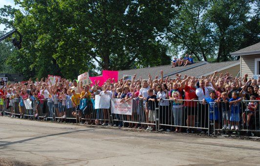 Large crowd behind a metal barricade, waving. Signs visible; sunny day. Building and trees in the background.
