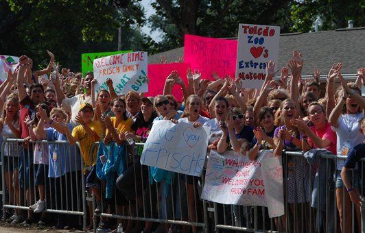 Crowd of people behind a metal barrier, waving and holding signs. Bright signs, cheers, and excitement visible.