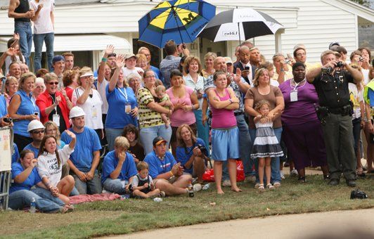 Large group of people gathered outdoors, some clapping, some holding umbrellas; a few wearing blue shirts.
