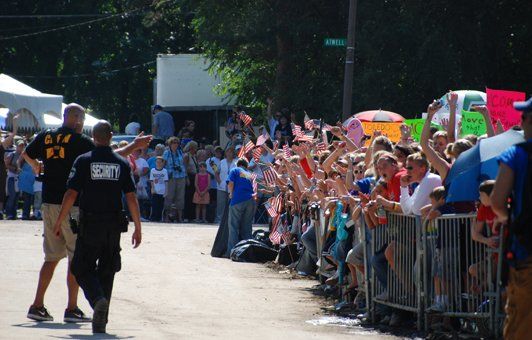 Crowd of people waving small flags behind metal barricades; security guards in foreground.
