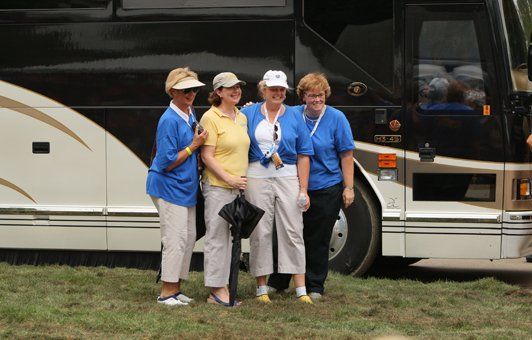 Four women in blue shirts and tan pants pose by a black tour bus on grass.