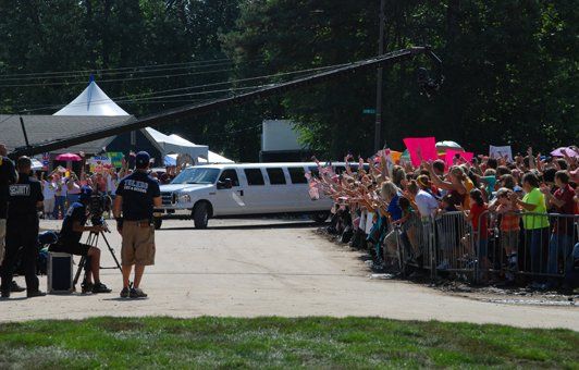 A white limousine drives down a road lined with a crowd of people waving. A boom mic is overhead.