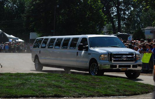 White limousine driving on grass, with crowd in the background.