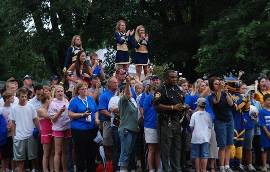 Crowd of people in blue and white, cheering. Cheerleaders atop shoulders. Police officer stands guard.