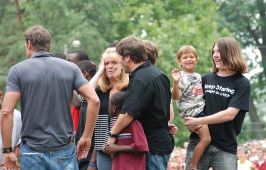 Group of people outdoors, some with children. Man in black shirt holds a boy, another waves. Others chat.