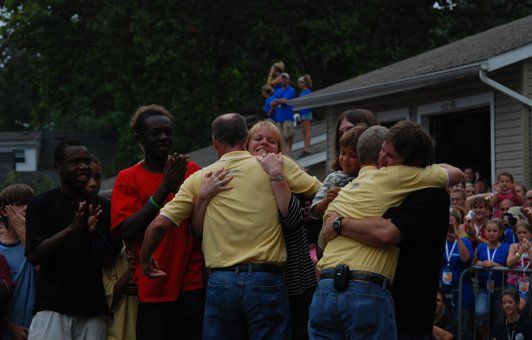 People embracing outdoors, some in yellow shirts. Group watching. Building in background.