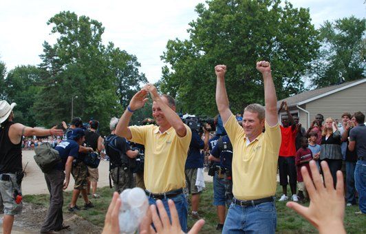 Two men in yellow shirts raise arms in celebration amidst a crowd outdoors.