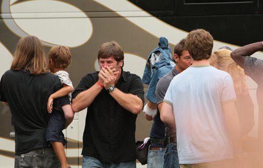 Man with hands over mouth, surrounded by people and a bus. A child is being held.