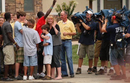 Family surrounded by news crew; woman cries, boy hugs her. Men carry cameras; outdoor setting.