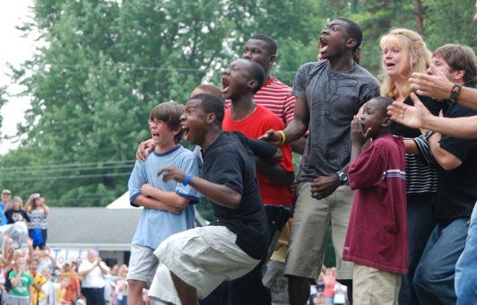 Group of people cheering outdoors, excited expressions, arms raised, green trees in background.