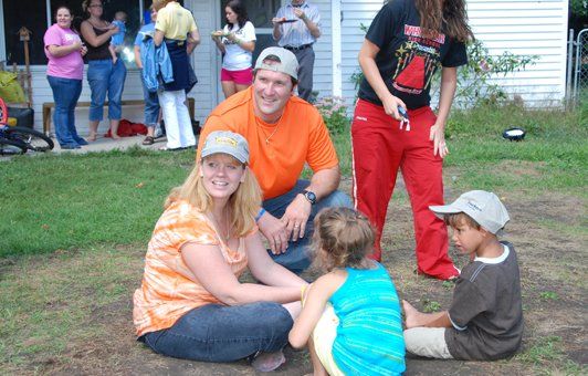 Family gathering outside; adults and children seated on ground. People in background near house.