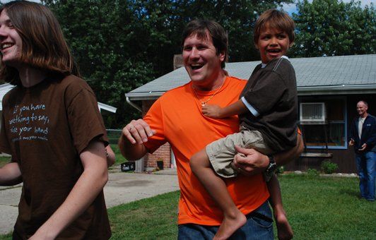 Man in orange shirt holds a child, smiles. Another person laughs nearby. Outside a house.