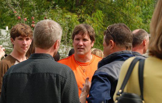 Men in a group, talking outside. One in orange shirt gestures, others listen. Green foliage in background.