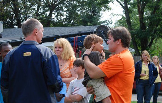 People gather outdoors; man in orange shirt holds a child, talking to another man. Others look on.