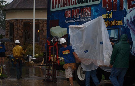 Delivery men unloading a large, plastic-wrapped appliance from a truck in the rain.