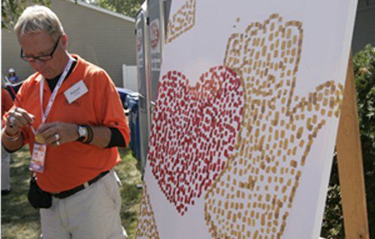 Man in orange shirt paints a heart and hand using small seeds on a canvas outdoors.