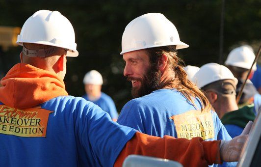 Men in blue shirts and white hard hats, talking outside. 