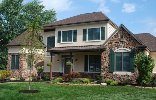 Two-story beige house with brown roof and stone facade accents, green shutters, and a well-manicured lawn.
