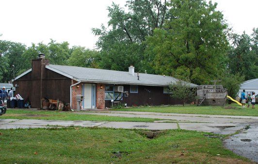 Brown house with a chimney and a small yard, trees in the background. A driveway and some people are visible.