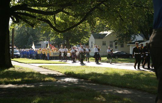 A marching band in kilts and formal wear parades down a street lined with houses on a sunny day.