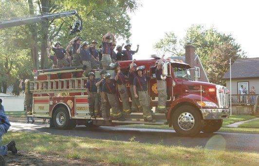 Firefighters on a red fire truck, waving. Outdoors in a residential area.