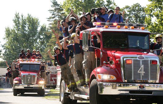 Firefighters ride atop two red fire trucks, waving. Trees and greenery in the background.