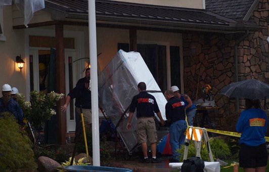 Emergency workers carrying a tarp-covered object near a house with a damaged entrance, possibly a collapsed structure.