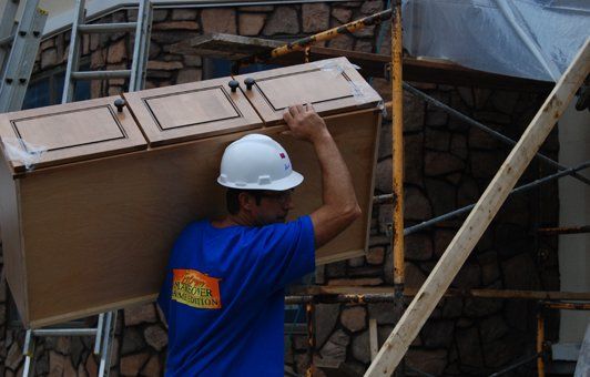 Construction worker carrying a brown cabinet outside, wearing a white hard hat and blue shirt.