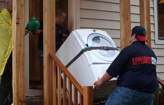Three people moving a white dryer down wooden stairs; the movers wear dark-colored shirts.