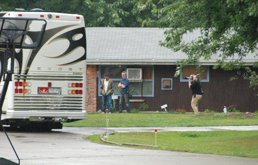 White RV parked near a brown house; people stand outside taking photos.