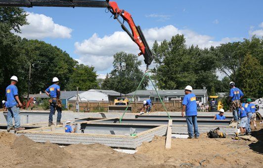 Construction workers lift modular wall with a crane. Men in blue shirts work on a foundation. Bright sunny day.