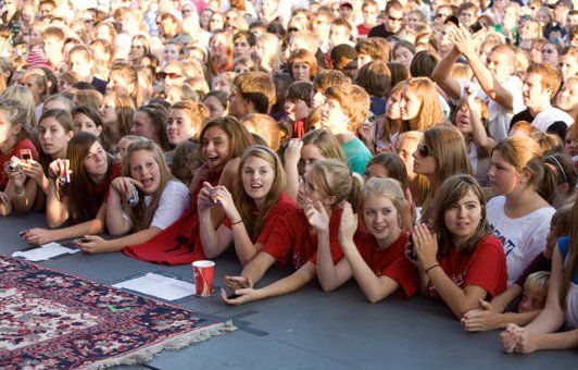 Large crowd of people gathered outdoors, many with long hair, some wearing red shirts, smiling and looking forward.