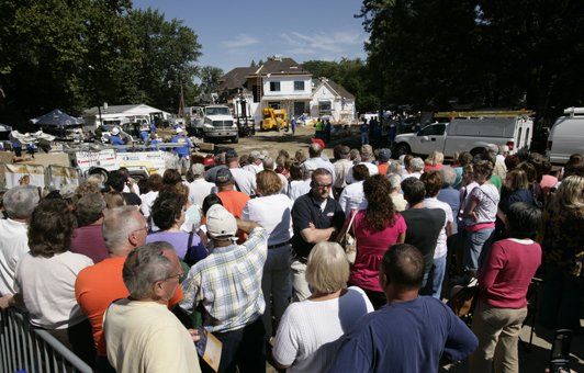 Large crowd gathered outdoors near a damaged house, possibly for an event or inspection.