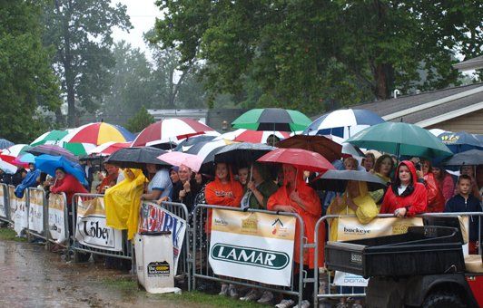 People under umbrellas watching an event in the rain, behind a barrier.