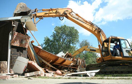 Yellow excavator demolishing a building, boat visible amongst debris, blue sky.