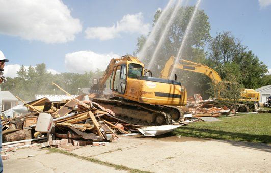 Yellow excavators demolishing a building, spraying water, with a worker in a hard hat nearby on a sunny day.