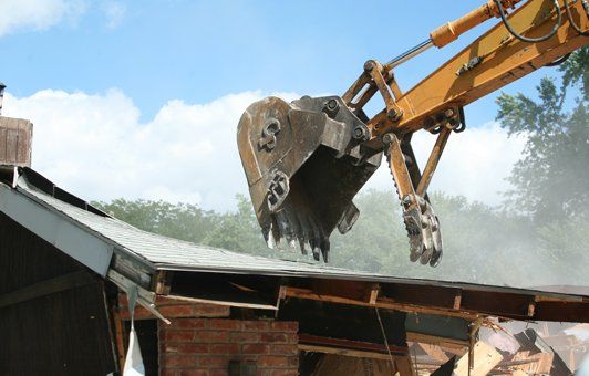An excavator demolishes a building with its bucket against a blue sky.
