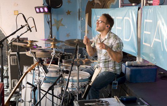 Drummer playing a light blue drum set on stage, looking up, light blue banner with white text in the background.
