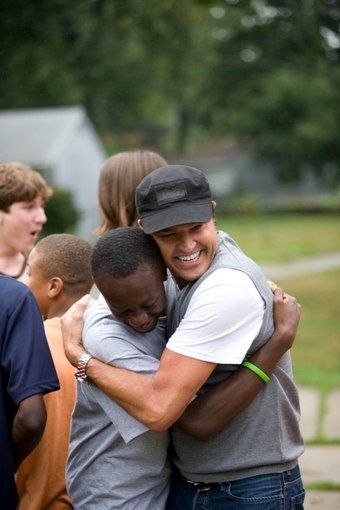 Two men embrace, smiling. Others in background. Outdoors, overcast day.