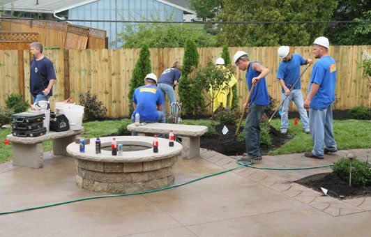 Workers in blue shirts and hard hats landscaping a backyard with fire pit and benches.